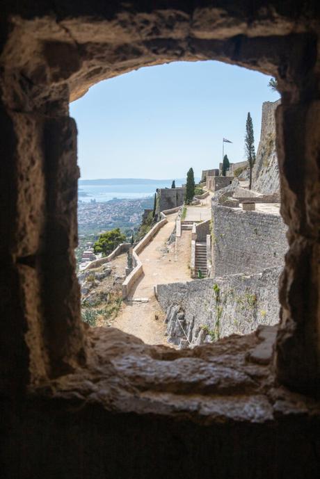 A view from within Klis Fortress through a stone window, offering a glimpse of the fortress walls and the expansive view of Split and the Adriatic coastline.