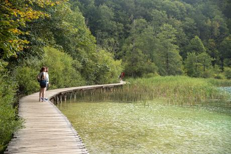 Two people stand on a wooden boardwalk that winds along a serene lake, surrounded by dense green forest. The peaceful path follows the edge of the water, blending into the natural environment.