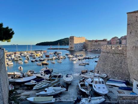 A picturesque harbor filled with small boats, enclosed by stone walls of a coastal fort, with a view of the open sea in the distance. The scene captures the calm waters and sunny blue sky.