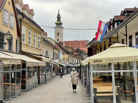 A quiet pedestrian street in the Croatian city of Zagreb, lined with outdoor cafes and small shops. A Croatian flag waves above the street, and a church steeple towers in the background.