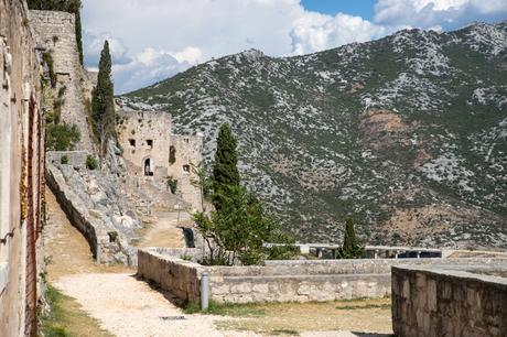 A close-up view of Klis Fortress in Croatia, with rugged stone walls blending into the surrounding rocky hills, a pathway winding up to an arched entrance.