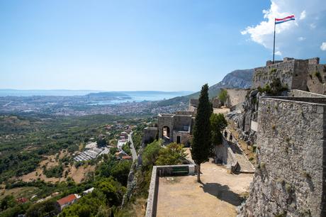 A sweeping view from Klis Fortress in Croatia, overlooking the city of Split and the Adriatic Sea, with a Croatian flag flying high in the wind.