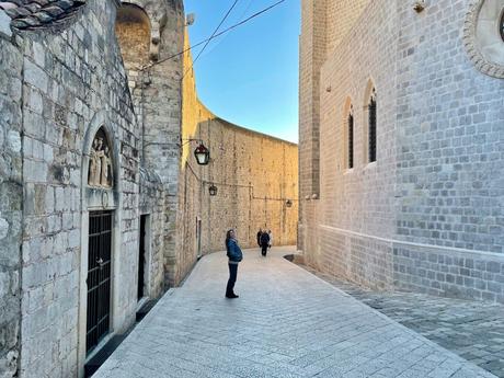 A narrow cobbled street lined with old stone buildings in the historic old town of Dubrovnik. A lone pedestrian stands in the middle, while a tall stone wall rises in the background.