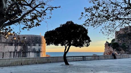 A twilight scene by the sea, featuring a large stone fortress illuminated by the setting sun. A lone tree stands silhouetted against the fading light, with the ocean in the background.