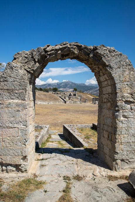 A view through a weathered stone archway, looking out towards ruins and distant mountains under a clear blue sky in Salona, Croatia.