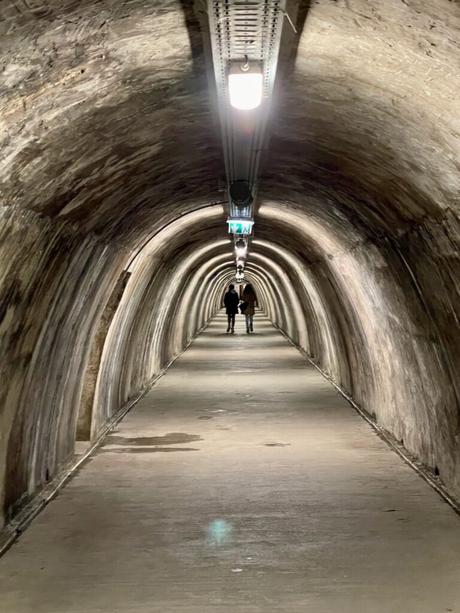 A narrow, dimly lit tunnel with an arched stone ceiling, leading into the distance. Two people walk toward the end of the tunnel, giving a sense of depth and mystery.