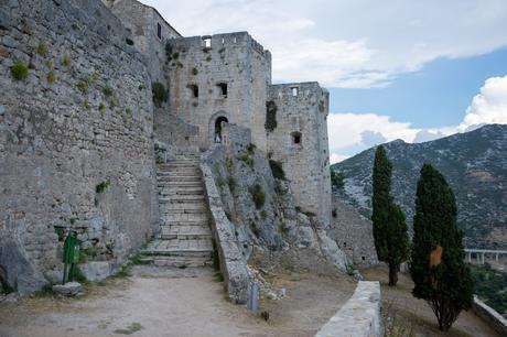 A path leading up stone steps to a towering entrance of Klis Fortress in Croatia, with steep stone walls on either side and the hills as a backdrop.