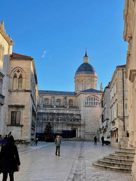 A cobblestone square in Dubrovnik with a few pedestrians, framed by tall historic buildings and a large domed church in the background. The bright blue sky contrasts with the stone architecture.