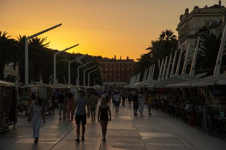 A busy promenade at sunset in Split, Croatia, with people walking and enjoying the market stalls under the orange and pink sky, palm trees lining the path.