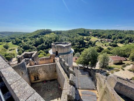 A panoramic view from the top of a castle ruin, looking out over green forests and rolling hills, with a section of the castle's round towers and walls in the foreground.