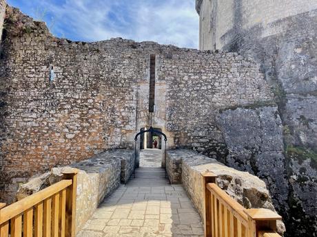 A stone bridge leading to the fortified walls of a medieval castle, with rugged stonework and a wooden handrail along the pathway.