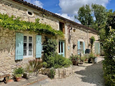 A quaint stone cottage in the south of France with pastel blue shutters, nestled in a small garden. The scene is bathed in sunlight, with lavender plants and green vines climbing the walls, creating a cozy and peaceful setting.