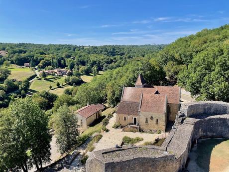 A picturesque view of a stone church nestled in a valley surrounded by lush green forests, with a small stone wall and medieval architecture visible.