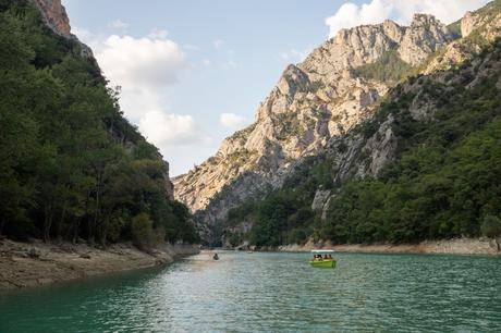 A calm river with turquoise water flowing between tall, rugged cliffs covered in trees. Small boats with people leisurely float along, surrounded by the grandeur of steep, rocky mountains.