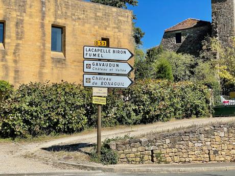 A rural road sign pointing to nearby villages such as Lacapelle Biron and Château de Bonaguil, against a stone wall and trees under a clear blue sky.