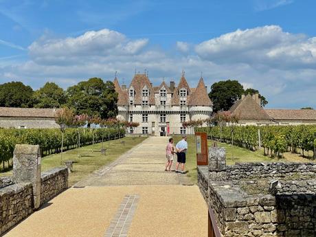 The grand Château de Monbazillac with multiple conical towers, framed by rows of grapevines and stone walls leading to its entrance, with two people admiring the view under a blue sky with scattered clouds.