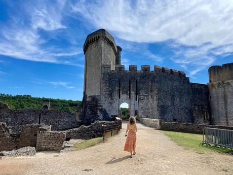 A woman in a flowing orange dress walks towards the entrance of a medieval stone castle, with towering walls and a blue sky overhead.