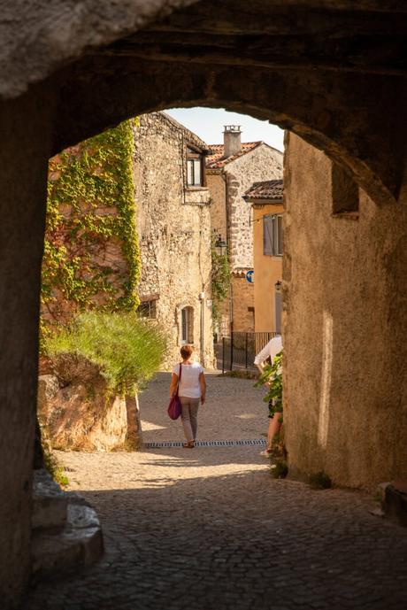 A cobblestone street in the Provencal village of Tourtour, framed by an archway made of stone. A woman with a pink bag is walking toward sunlit stone houses adorned with ivy, creating a peaceful, timeless atmosphere.