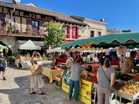 A traditional outdoor market with stands selling fresh produce like tomatoes and zucchinis, and people browsing under the shaded stalls in a sunny French town.