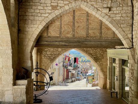 A medieval stone archway in a French village, looking out toward a street decorated with vibrant flags, with shops and buildings visible in the distance.