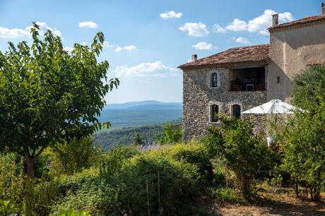 A charming stone house in Provence with a red-tiled roof surrounded by lush greenery. A white patio umbrella is set up outside, and the view stretches out to rolling hills and mountains under a blue sky with scattered clouds.