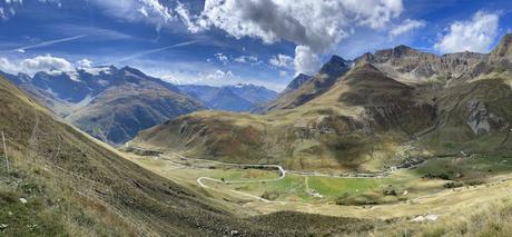 A panoramic view of a vast alpine landscape with rolling hills and winding roads stretching across green valleys, framed by tall mountain peaks in the distance.