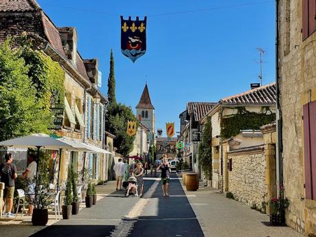 A lively town street with banners hanging above, where people stroll past historic stone buildings and an old church tower, basking in bright, sunny weather.