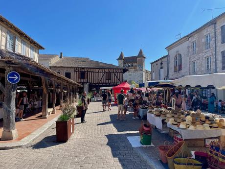 A vibrant town market scene in Villereal, France with multiple stalls displaying a variety of goods, and people shopping in the foreground, set against historic half-timbered buildings.