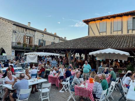 A popular outdoor dining area with people seated at tables covered in red and white checkered tablecloths. The scene is set in a historic town square, with a traditional market structure and old stone buildings in the background.