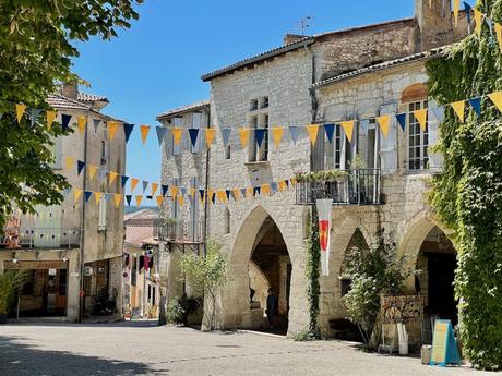 A village square with stone buildings, arched walkways, and yellow and blue bunting strung overhead, creating a festive atmosphere under a bright blue sky.