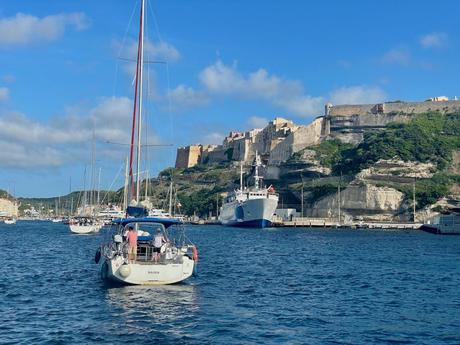 A marina with sailboats docked in the harbor, overlooked by a large, historic fortress perched on a rocky cliff, with the blue sky and sea adding to the peaceful atmosphere.