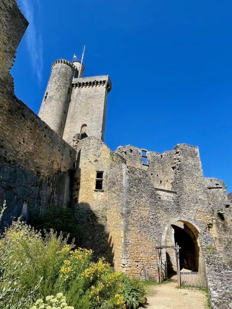 A view of a tall stone castle tower against a bright blue sky, with ancient stone walls and wildflowers growing at the base.