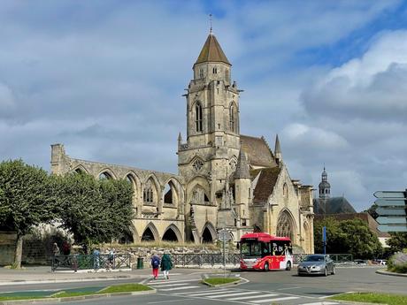 A picturesque view of a historic stone church in Caen with Gothic arches, surrounded by trees and a small road where a red bus and a car are passing by. The structure shows a mix of intact and partially ruined sections, standing under a bright blue sky.