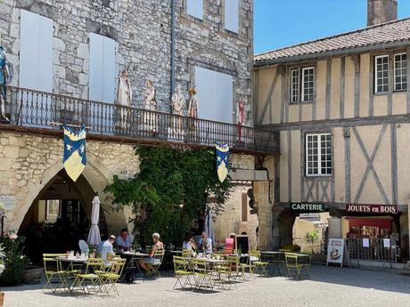 A café scene in a charming French village with customers seated at small tables under the arches of a historic building, with decorative flags and stone architecture.