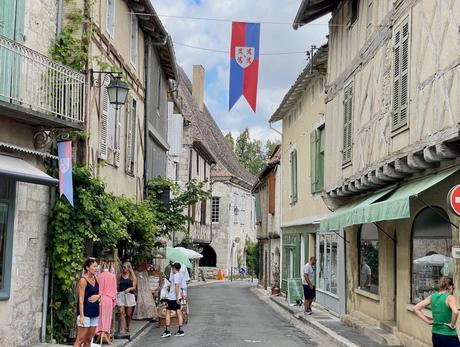 A peaceful medieval street lined with timber-framed houses, where tourists browse outdoor shops, under a vibrant flag hanging above the road.