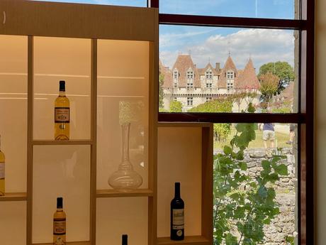 A view of the château through a modern window inside a wine shop, showcasing several bottles of wine on display while the château and its surroundings are visible in the distance.