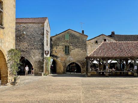 An open square in the medieval town of Monpazier with an arched stone market building, dotted with tourists exploring its cobbled pathways under a bright blue sky.