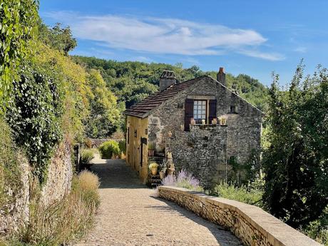 A charming stone house with a weathered exterior sits along a narrow cobblestone path, surrounded by vibrant greenery under a clear blue sky.