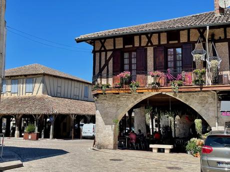 The charming Villereal town square featuring stone buildings with wooden beams and flower boxes. A small café sits under the archways, and a sunny blue sky adds to the inviting atmosphere of the quiet town.