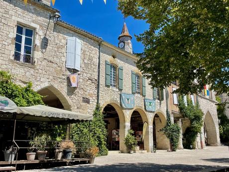 A picturesque square with a clock tower and stone arches, surrounded by leafy trees and a café with an awning that reads 