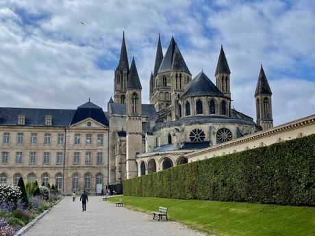 The grand stone abbey in Caen with towering spires and arched windows stands prominently against a partly cloudy sky. The foreground features a neatly manicured garden and pathway leading toward the building.