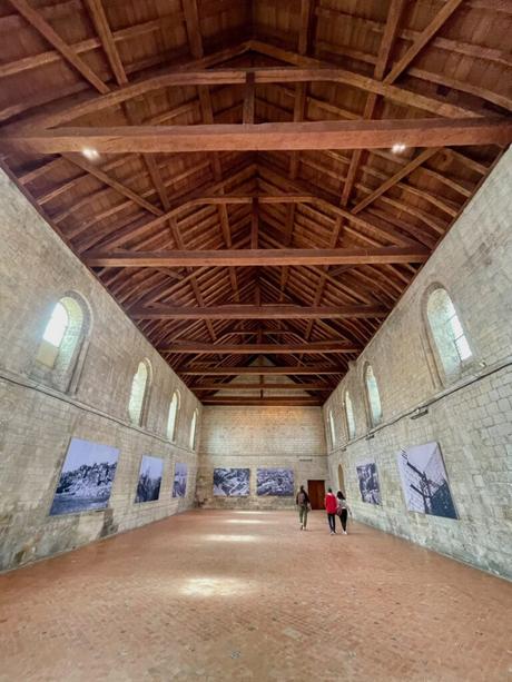 A spacious, rustic hall with exposed wooden beams on the ceiling. The stone walls are lined with black-and-white photographs, and two visitors walk towards the far end of the room, giving the space a contemplative feel.