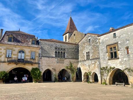 A quaint town square featuring arched buildings with wooden shutters, framed by trees and a towering old church with a red-tiled roof.