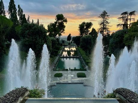 A grand garden scene in Tivoli's Villa d'Este at sunset, featuring tall water fountains in the foreground, perfectly aligned rectangular pools, and well-manicured hedges leading into the distance, with a backdrop of trees and a colorful sky.