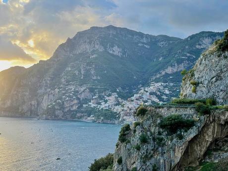 A closer view of a steep coastal mountain covered with green trees, leading down to a small, densely packed town built along the cliffside with white buildings, bathed in soft sunlight.