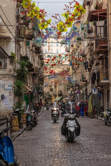 A narrow street in Naples, decorated with festive hanging lights, where people ride motorbikes along cobblestone roads surrounded by historic buildings.