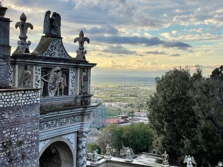 An ornately carved stone archway featuring decorative sculptures of figures and symbols, overlooking a distant valley with a dramatic cloudy sky as the sun begins to set.