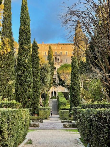 A formal garden with tall, slender cypress trees lining a walkway leading up to a grand villa in the distance. The garden features neatly trimmed hedges and a golden light hitting the scene.