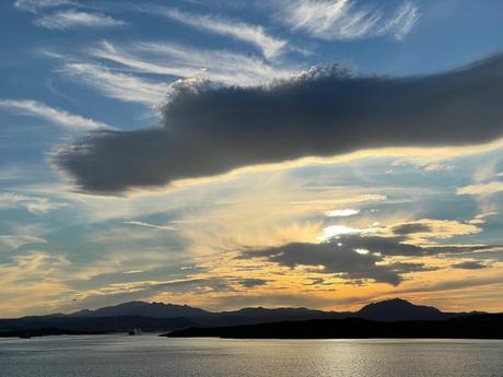 A serene sunset over a calm sea with dramatic clouds in the sky, casting a warm glow over distant mountains and the horizon.