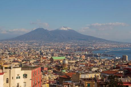 A panoramic view of Naples, Italy, with Mount Vesuvius in the background, overlooking the bustling city and its harbor.